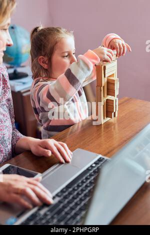 Woman mother working doing her job remotely during video chat call stream online course webinar on laptop from home while her daughter playing with br Stock Photo