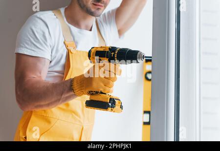 Handyman in yellow uniform installs new window by using automatic screwdriver. House renovation conception Stock Photo