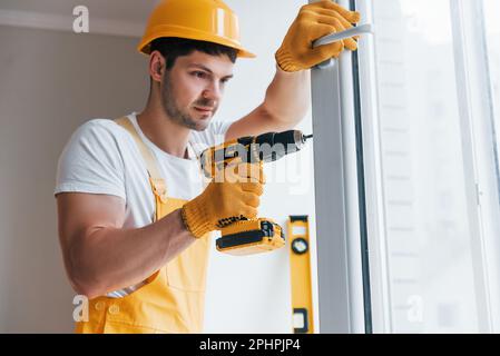 Handyman in yellow uniform installs new window by using automatic screwdriver. House renovation conception Stock Photo