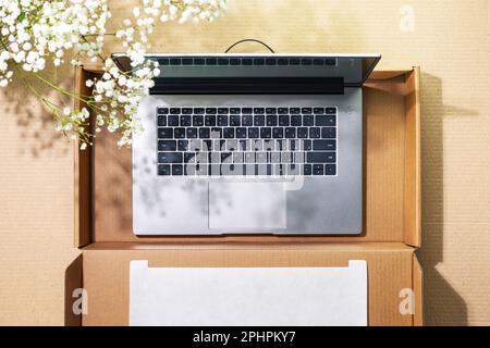 Buying and delivering new technics. Romantic sustainable overhead shot of laptop in cardboard box on cardboard background with white spring flowers an Stock Photo
