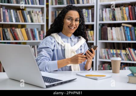 Portrait of a beautiful Hispanic female student in a university library among books, a woman with curly hair is smiling and looking at camera, holding hone, using an application online learning. Stock Photo