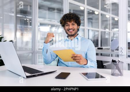 A young man, an Indian student, received good news by letter, he entered the university, passed the exam, and got a job. Sitting in the office, happy, smiling, looking at the camera. Stock Photo