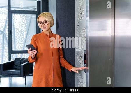 A smiling young business woman stands in the office center near the elevator, presses the button, makes a call. He holds the phone in his hands, reads messages, dials, calls. Stock Photo