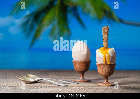 Soft boiled egg in eggcup with slice of toasted toast on wooden table with sea water, coconut palm tree and blue sky background on sunny summer day in Stock Photo