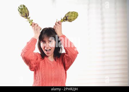 Joyful young Caucasian woman holding two artichokes as horns on her head, laughing and looking at the camera, standing by a window with a white curtai Stock Photo