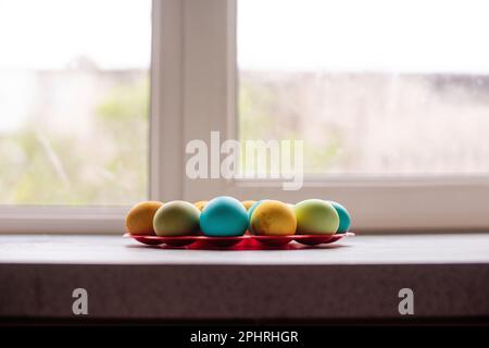 On a red, round stand near the window, bright Easter eggs lie on a marble table. Homogeneous lifestyle background. Easter traditions to paint eggs. Be Stock Photo