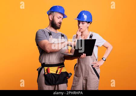 Renovators team holding laptop and browsing internet online, discussing about renovation project. Construction workers using wireless pc to search building inspiration, studio shot. Stock Photo