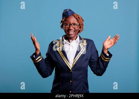 Professional female flight attendant happily greeting arriving passengers at airplane cabin. Woman wearing stewardess uniform looking at camera in studio shot against blue background. Stock Photo