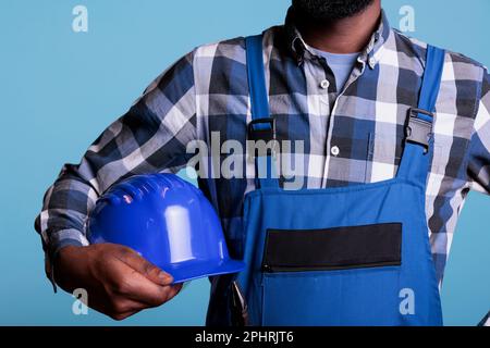 Middle section of construction worker holding protective helmet under his arm. African American man wearing coverall and plaid shirt against blue background, studio shot. Stock Photo