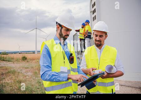 group of wind mill professional engineers contractors working and using a tablet to watch the draft of the construction of the generator propeller of renewable energy wearing helmet and safety vest. High quality photo Stock Photo