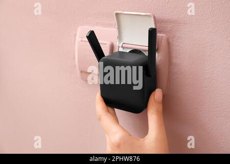 Woman plugging black WiFi repeater in electric socket on pink wall Stock Photo