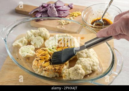 Hand with a silicone brush spreading spice oil on raw cauliflower slices in a glass casserole, cooking preparation for baked vegetable steaks, selecte Stock Photo