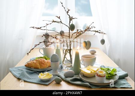 Breakfast table with hanging Easter eggs on spring branches, candles, and blue gray decoration in front of a window with white curtains, copy space, s Stock Photo