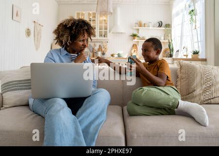 African American boy kid asking attention from busy mother freelancer while sitting on sofa at home Stock Photo