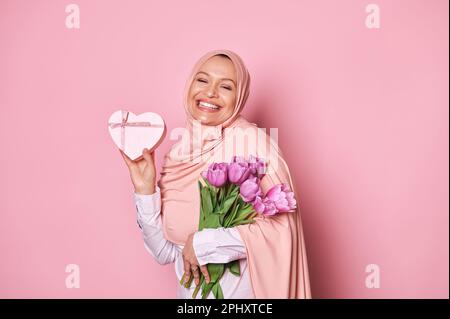 Overjoyed Arab Muslim woman in pink hijab, smiling looking at camera, posing with gift box and bouquet of purple tulips Stock Photo
