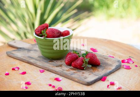 Wooden board with green bowl strawberries lying on table with roses petals outdoor. Stock Photo