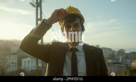 Ambitious young urban planner or businessman in stylish suit and helmet touches his helmet and smiles proudly against cityscape background Stock Photo