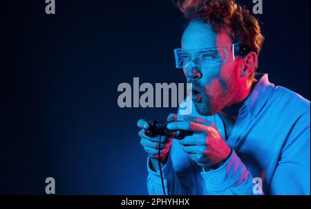 Plays game by using controller. Neon lighting. Young european man is in the dark studio Stock Photo