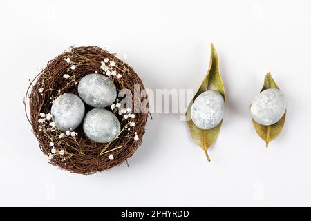 Brown nest of twigs with three gray Easter eggs, feathers, dried magnolia leaves and sprigs of gypsophila on gray background. Minimalistic Easter conc Stock Photo