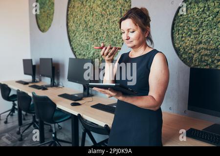 Businesswoman having business call working on tablet in office. Mature busy woman using touch pad computer standing by window in modern interior. Fema Stock Photo
