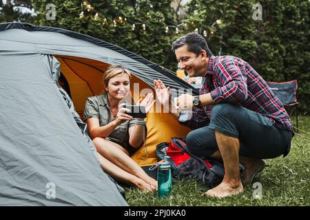 Couple having video call with friends using smartphone while sitting in tent at camping. People relaxing in tent during summer vacation. Actively spen Stock Photo