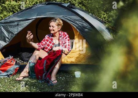 Female having video call with friends using smartphone while sitting in tent at camping. Woman relaxing in tent during summer vacation. Actively spend Stock Photo