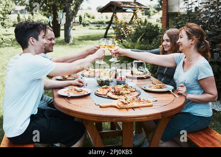Friends making toast during summer picnic outdoor dinner in a home garden. Close up of people holding wine glasses with white wine over table with piz Stock Photo