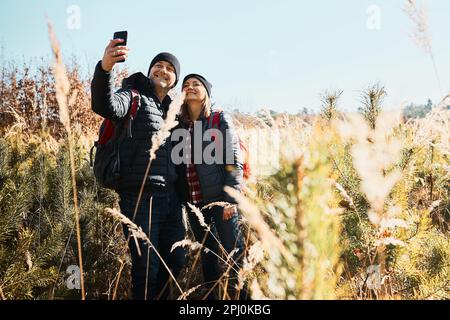 Hugging couple taking selfie while vacation trip. Hikers with backpacks on way to mountains. People walking through tall grass along path in meadow on Stock Photo