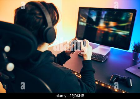 Man playing video game at home. Gamer holding gamepads sitting at front of screen. Streamer playing online in dark room lit by neon lights. Competitio Stock Photo