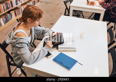 Student learning in university library. Young woman writing essay and making notes using computer tablet. Focused student studying for college exams. Stock Photo