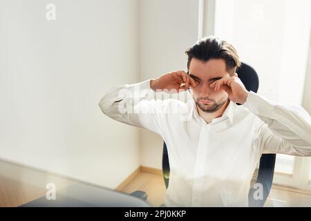 Tired exhausted young man rubbing eyes feeling eyestrain from long computer work. Man wearing white shirt sitting at computer screen Stock Photo