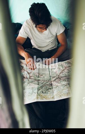 Young man looking at a map of mountain trails planning next trip on summer vacation sitting in a tent on camping. Concept of camp life Stock Photo