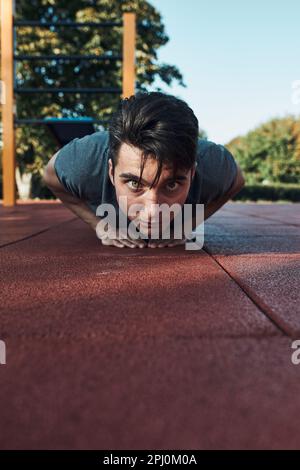 Young man doing push-ups on a red rubber ground during his workout in a modern calisthenics street workout park. Man wearing sportswear looking at cam Stock Photo