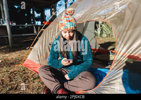 Young traveler woman sitting in the door of a camping tent and using her mobile phone at the morning Stock Photo