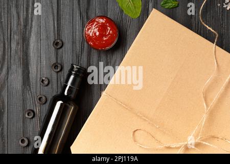 Pizza in a closed cardboard box, sauces, olive oil with spices, fresh vegetables on a dark wooden background top view. Food delivery. Stock Photo