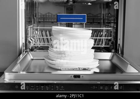 Built-in dishwasher with open door and clean dishes. Clean white plates sit on the open door of the dishwasher. Household appliances for the kitchen. Stock Photo