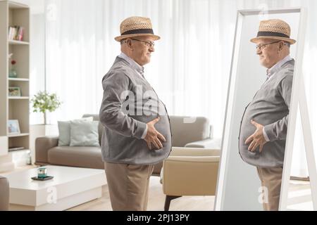 Elderly man with a big belly looking at a mirror at home in a living room Stock Photo