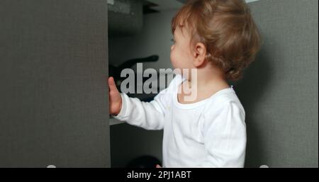 One year old baby boy opening kitchen cabinet cupboard closer door. Infant toddler standing at kitchen floor Stock Photo