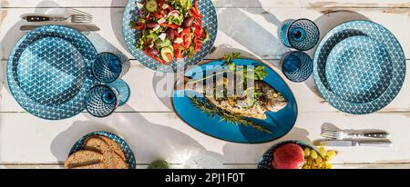 Gilt-head bream also known as Orata baked in the oven and served on a white wooden table with vegetable salad and fruits. Healthy seafood preparation Stock Photo
