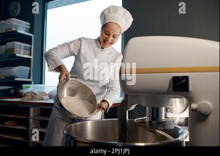 Woman baker pouring flour in mixing machine at bakery Stock Photo