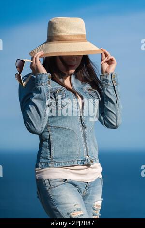 Portrait of hipster woman in denim jacket and jeans hiding with straw hat over his face. Adult female posing on background of blue sky and ocean Stock Photo