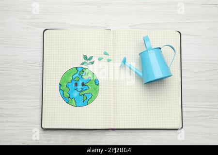 Happy Earth Day. Notebook with drawing of planet and watering can on white wooden table, top view Stock Photo