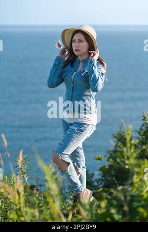 Full length portrait woman dressed in straw hat, denim jacket, jeans and boots standing on seashore in sunny weather. Hipster female raised hands Stock Photo