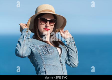 Caucasian ethnicity woman in sunglasses raised her hands and holding straw hat on her head. 40 year old hipster woman in denim jacket standing Stock Photo