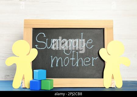 Small chalkboard with phrase Surrogate Mother, paper people cutouts and cubes on blue wooden table Stock Photo