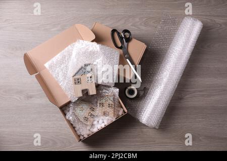 Open box with decorative house figures, adhesive tape, scissors, foam peanuts and bubble wrap on wooden table, flat lay Stock Photo