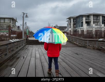 Woman under rainbow umbrella. Person walking with colorful umbrella on the pier at rainy day. Woman holding multicolor umbrella in overcast day. Stree Stock Photo