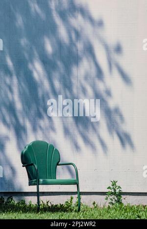 Vintage green metal scallop shell chair on lawn in front of white sun-washed wall with patterned shadow of foliage. Stock Photo