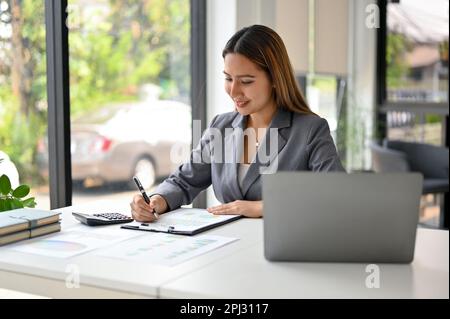 Smiling and attractive millennial Asian businesswoman examining and analyzing sales data on reports at her desk. Stock Photo