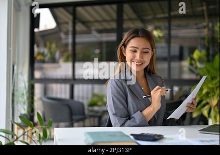 Attractive millennial Asian businesswoman or female financial worker analyzing financial data on reports and working at her desk. Stock Photo